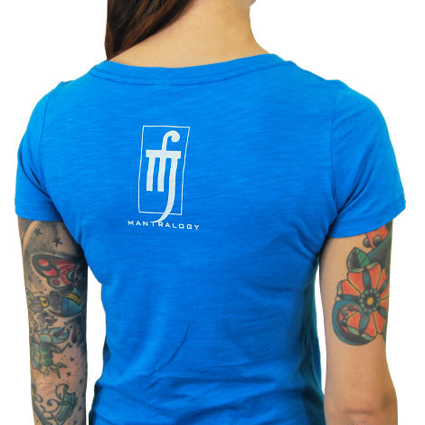 A person with colorful arm tattoos wears a bright blue Mantralogy T-shirt with a white stylized logo and Mantralogy printed below it on the back, set against a plain white background.