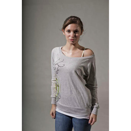 A young woman with light brown hair wears a loose Mantralogy Shantala T-shirt featuring a Saraswati-inspired print, blue jeans, and a visible white camisole strap, standing facing forward against a plain gray background.