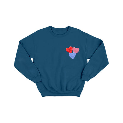 The glimmers blue crewneck sweatshirt features three candy hearts on the left chest—two red and one purple—with text styled like conversation heart candies.