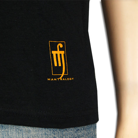A close-up of a black Mantralogy shirt with a small yellow logo near the hem, next to a mannequin’s arm, and part of blue jeans visible below.