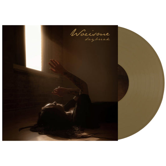 The Woe Is Me Daybreak Gold Vinyl features cover art of a person lying on a wooden floor, reaching for sunlight through a window, gold script album title at top right. Includes a full album digital download presale.