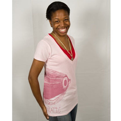 A smiling woman with short hair and a gold necklace stands before a white background, wearing a Mantralogy light pink T-shirt with a large pink fish graphic on the side over a red V-neck shirt.