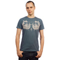 A young man with short hair faces the camera, wearing a Mantralogy blue graphic T-shirt with white designs and dark jeans, set against a plain white background.