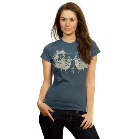 A woman with long brown hair wears a Mantralogy blue graphic t-shirt and dark jeans, standing against a white background with one hand in her pocket and a relaxed expression.