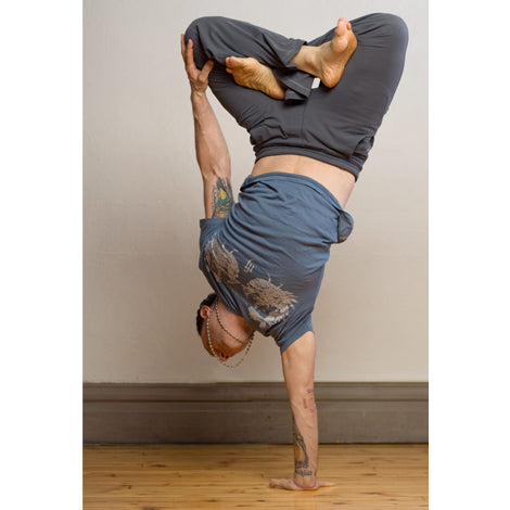 A person practices a one-hand yoga pose on a wooden floor against a light wall, wearing Mantralogy attire: blue shirt, gray pants, and a beaded necklace, with legs bent and crossed.