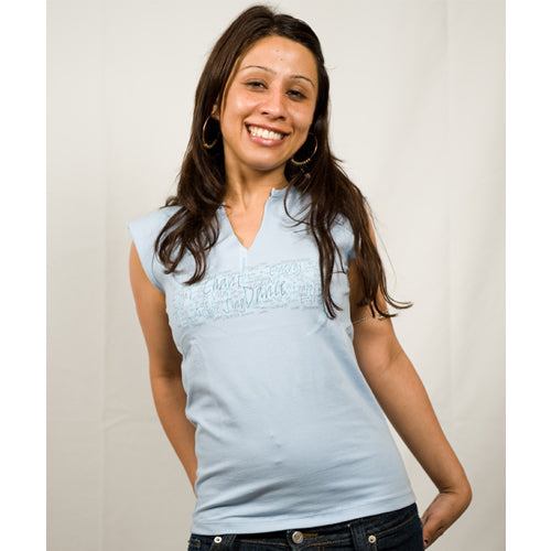 A woman with long brown hair beams joyfully in a light blue sleeveless Mantralogy top and jeans, standing against a plain light background with her hands behind her back, radiating the bliss of uplifting experiences.