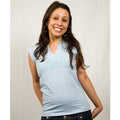 A woman with long brown hair beams joyfully in a light blue sleeveless Mantralogy top and jeans, standing against a plain light background with her hands behind her back, radiating the bliss of uplifting experiences.