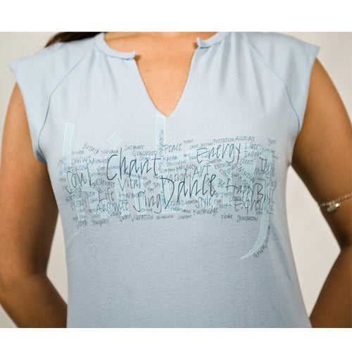 A person wears a Mantralogy light blue top with a notched neckline, featuring Chant, Sing, and Dance in mixed fonts inspired by kirtan music across the chest. Their arms are partly visible.