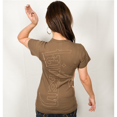 A woman with long brown hair stands facing away, wearing a Mantralogy brown lifestyle t-shirt with large vertical text on the back. She raises her left arm, bent at the elbow, against a plain white background.