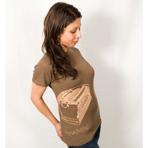 A woman with long brown hair wears a Mantralogy brown t-shirt with a harmonium graphic and the word HARMONIUM, standing against a white background—an ideal shirt for Kirtan music lovers.