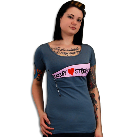 A tattooed individual wears a blue Mantralogy T-shirt featuring a pink and white graphic of an unzipped section revealing Occupy ♥ Street, posed against a plain white background.