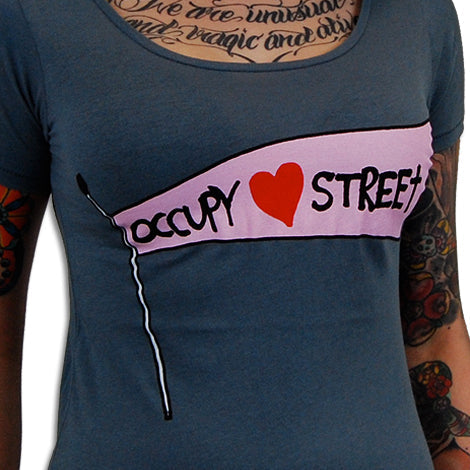 A person in a blue Mantralogy shirt featuring a pink Occupy ♥ Street flag graphic, showing colorful tattoos on their chest and arms.