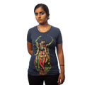 A woman with a serious expression stands against a white background, wearing a blue organic cotton Sadbhuj shirt from Mantralogy/Gaura Vani. The shirt features a vibrant artistic image of Sri Chaitanya playing a flute. Her hands are clasped behind her back.