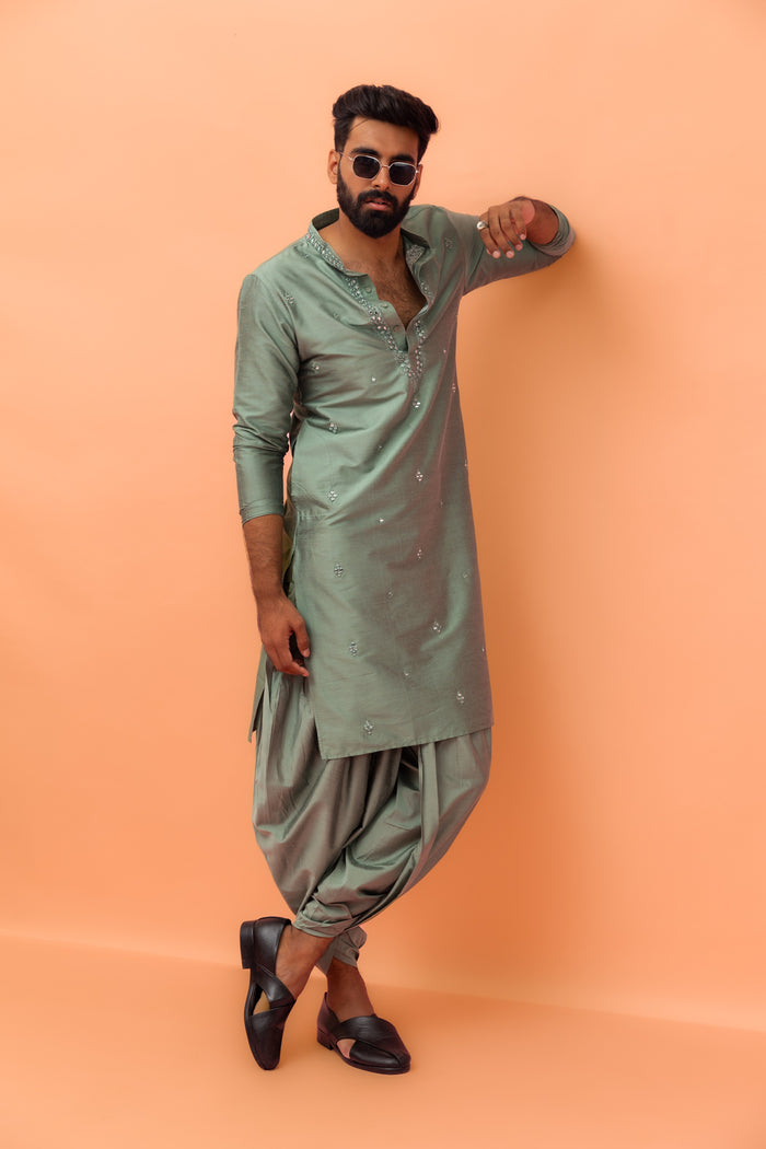 The model in sunglasses is leaning against the wall, posing in Kisah Sage Green Mirror Work Kurta with Dhoti set