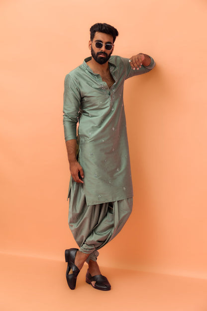 The model in sunglasses is leaning against the wall, posing in Kisah Sage Green Mirror Work Kurta with Dhoti set