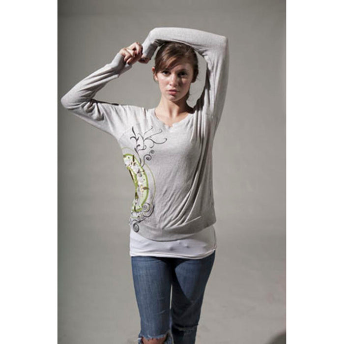 A woman wears a light gray Mantralogy Shantala T-shirt, featuring a decorative Saraswati-inspired design, paired with ripped blue jeans. She poses with one arm raised against a plain, neutral background.