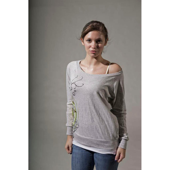 A young woman with light brown hair wears a loose Mantralogy Shantala T-shirt featuring a Saraswati-inspired print, blue jeans, and a visible white camisole strap, standing facing forward against a plain gray background.