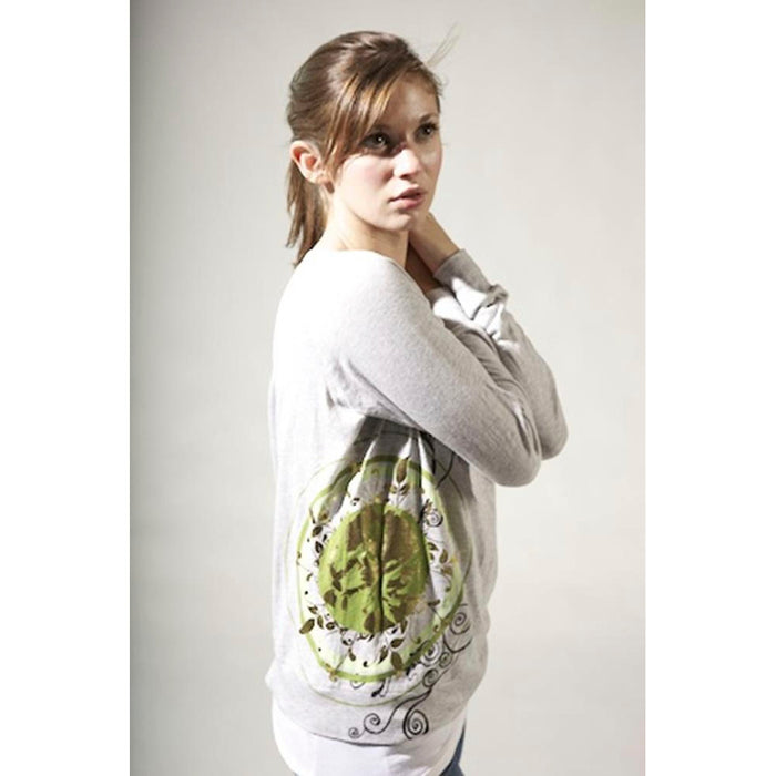 A young woman with brown hair in a ponytail wears a light gray Mantralogy Shantala T-shirt featuring a green and yellow circular Saraswati-inspired design, looking over her shoulder against a plain light background.