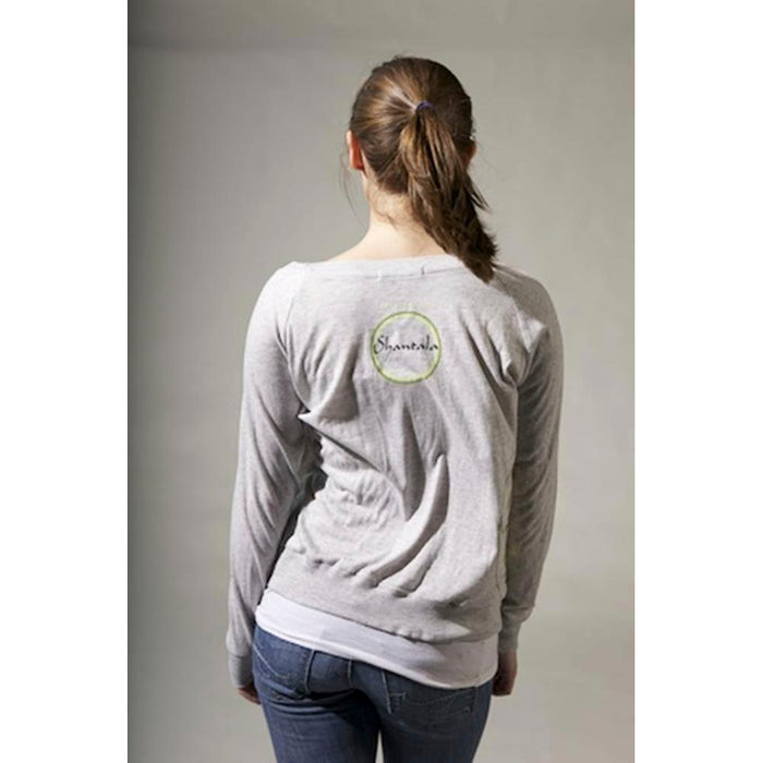 A woman with brown hair in a ponytail faces away, wearing a light gray Mantralogy Shantala T-shirt and blue jeans. The shirt’s back has a small circular logo and Shareable, inspired by Saraswati, Goddess of art and music.