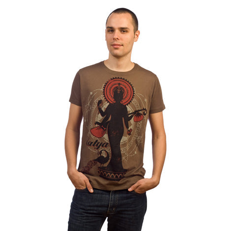 A person wearing a Mantralogy brown organic cotton T-shirt featuring a large graphic of a four-armed Indian deity and the word satya, standing with hands in pockets against a white background.