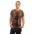 A person wearing a Mantralogy brown organic cotton T-shirt featuring a large graphic of a four-armed Indian deity and the word satya, standing with hands in pockets against a white background.