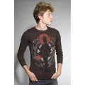 A young man with light brown hair wears a Mantralogy dark long-sleeve shirt featuring a bold red and black deity graphic, stands with hands in pockets, and looks to his left against a plain background.