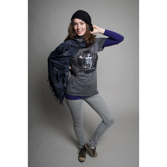 A woman smiles against a gray background, wearing an ultra-soft Mantralogy burnout shirt, black knit hat, dark scarf, light gray jeans, and brown shoes—one hand on her hat and the other holding her scarf.